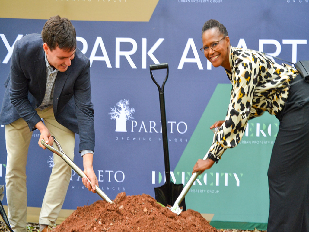 Two people in business attire smile whilst holding spades and digging into a pile of soil at the groundbreaking ceremony for the Pareto Menlyn Park development, with company banners in the background. - Pareto Properties