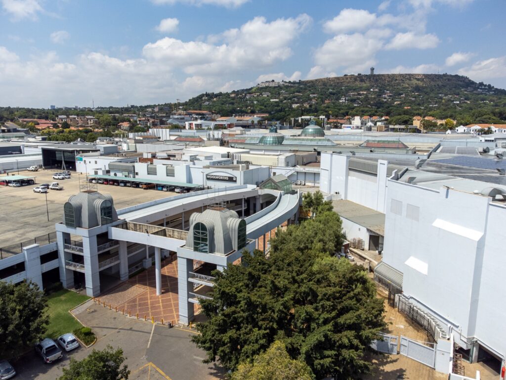 A large shopping centre complex, Cresta Shopping Centre, with multiple entrances, car parks, and surrounding trees, set against a backdrop of hills and a partly cloudy sky. - Pareto Properties