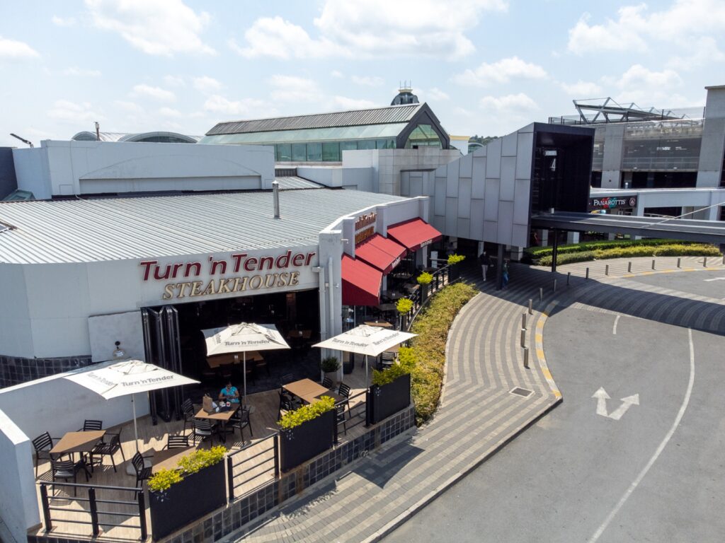 Outdoor seating area of Turn 'n Tender Steakhouse at Cresta Shopping Centre, with parasols, tables, and nearby modern buildings under a partly cloudy sky. - Pareto Properties