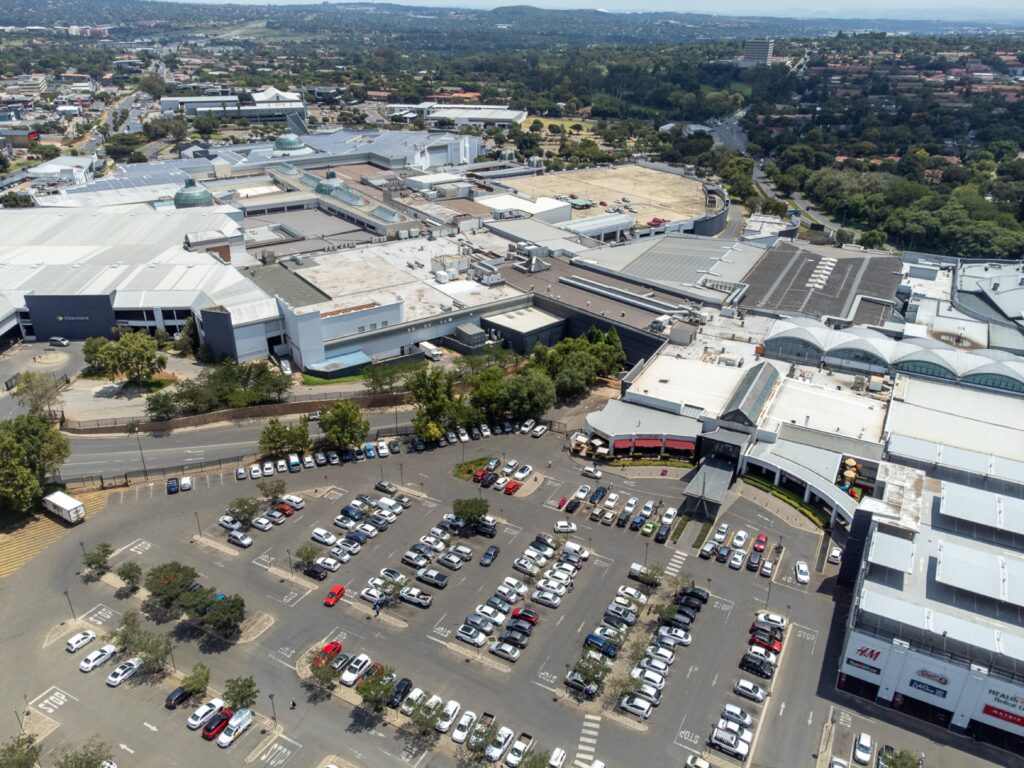 Aerial view of Cresta Shopping Centre, a large shopping centre complex with a busy outdoor car park and several cars, surrounded by trees and urban landscape. - Pareto Properties