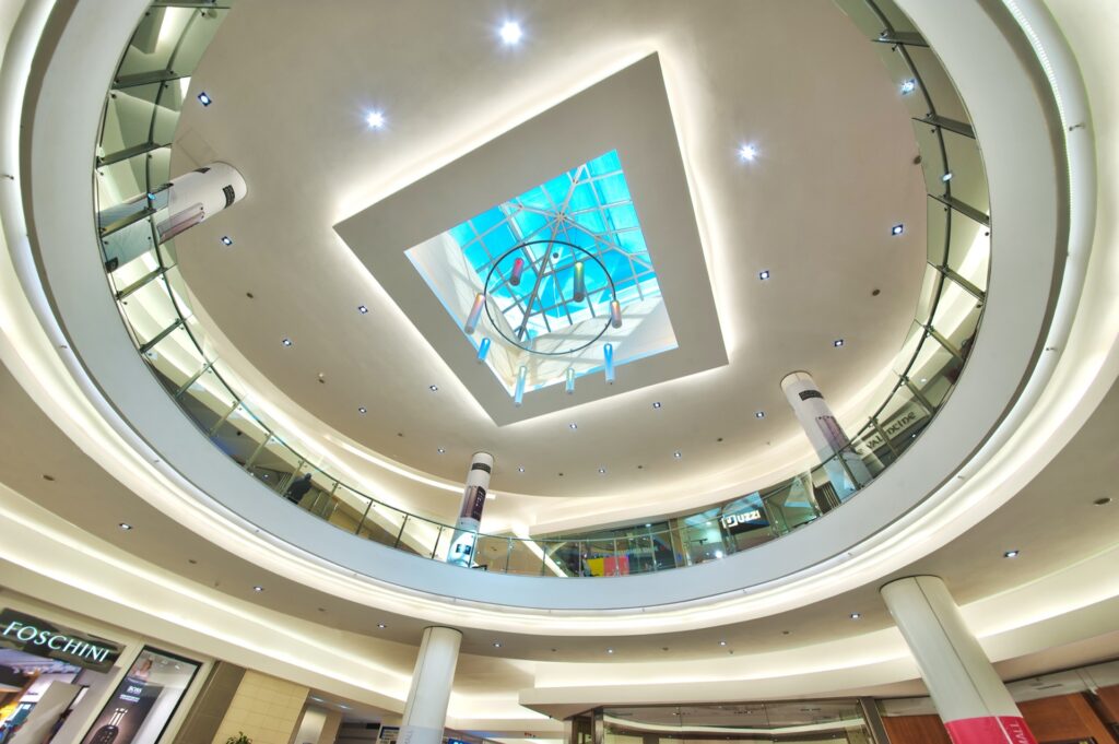 View of Mimosa Mall Bloemfonteinâ€™s modern shopping centre interior with circular balconies, bright lighting, glass railings, and a skylight ceiling featuring a geometric design and a hanging sculpture. - Pareto Properties