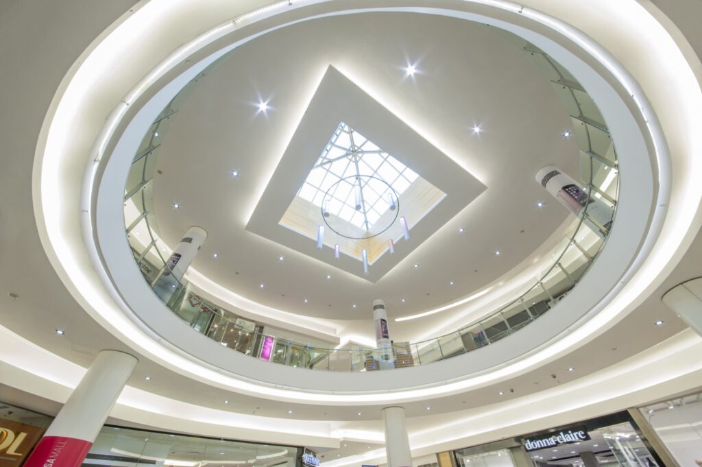 View looking up at Mimosa Mall Bloemfonteinâ€™s modern shopping centre circular atrium, featuring glass railings, multiple floors, bright lighting, and a geometric skylight at the ceilingâ€™s centre. - Pareto Properties