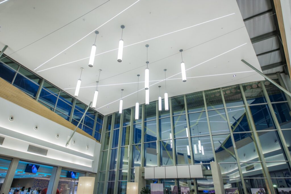 Modern building interior with large glass windows, high ceiling, and multiple hanging lights. LED light strips are embedded in the white ceiling, creating geometric patterns reminiscent of the contemporary design found at Mimosa Mall Bloemfontein. - Pareto Properties