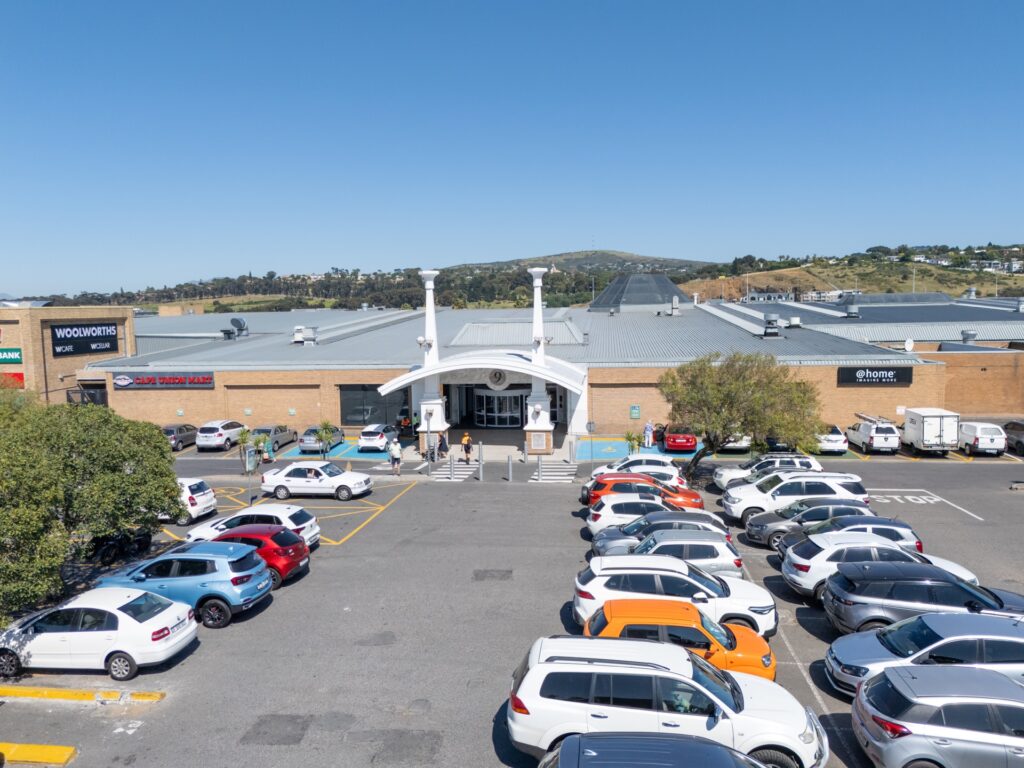 A shopping centre with multiple shops, a car park filled with cars, and a clear blue sky in the background, reminiscent of Tyger Valley Centre Bellville. - Pareto Properties