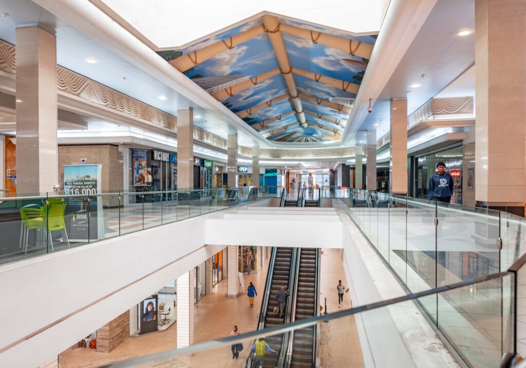 The interior of Westgate Mall Roodepoort features modern escalators, sleek glass railings, a beautifully painted ceiling, and several people walking and shopping throughout the vibrant space. - Pareto Properties
