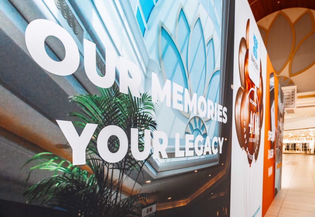 Large wall display at Westgate Mall Roodepoort with bold white text reading "OUR MEMORIES YOUR LEGACY" against a backdrop of an indoor architectural scene and a potted plant. - Pareto Properties