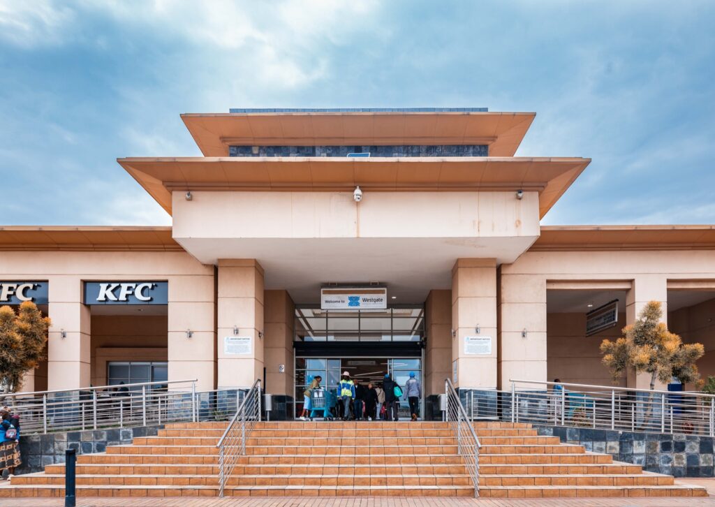 Wide view of a modern building entrance at Westgate Mall Roodepoort, with steps leading up, people entering, and KFC signs visible on both sides. - Pareto Properties