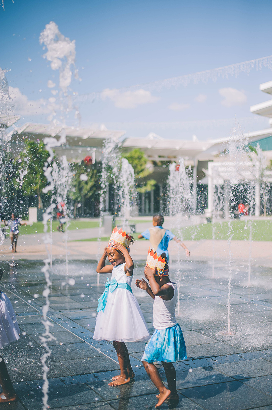 Two children wearing paper crowns play in a water fountain area outdoors on a sunny day, with buildings and more children visible in the background. - Pareto Properties