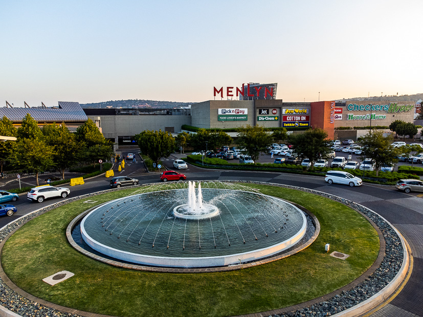 A large circular fountain with water jets in front of Menlyn Park Shopping Centre, with visible shop signs and parked cars in the background. - Pareto Properties