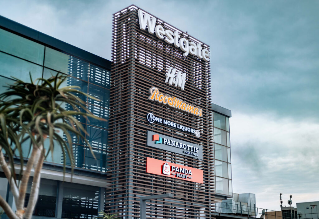 Shopping centre exterior with a tall sign displaying logos for Westgate, RoccaMamas, One More Liquor, Panarottis, and Panda, against a cloudy sky. - Pareto Properties