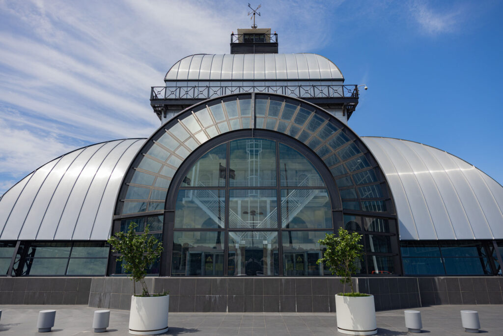 A modern building with a large arched glass entrance, metallic curved roof, and potted plants on a paved area under a blue sky. - Pareto Properties