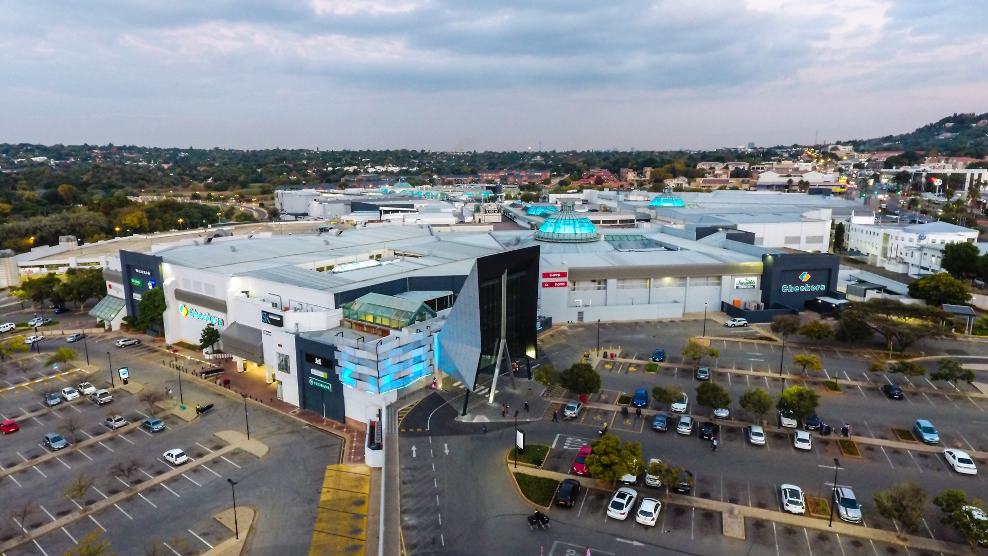Aerial view of a large shopping centre, part of the Pareto portfolio shopping centres, with a mostly empty car park, modern architecture, and surrounding trees and buildings under a cloudy sky. - Pareto Properties