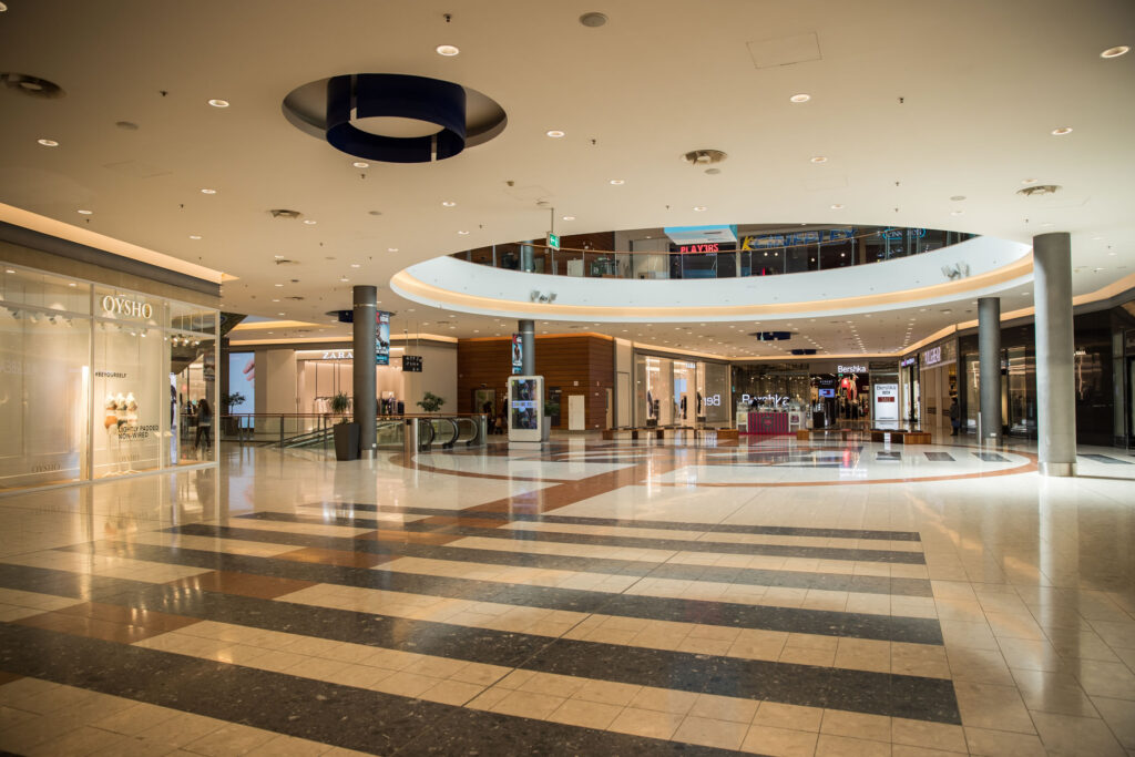 Interior of a modern shopping centre with bright lighting, empty walkways, various shopfronts, and a lift in the background. - Pareto Properties