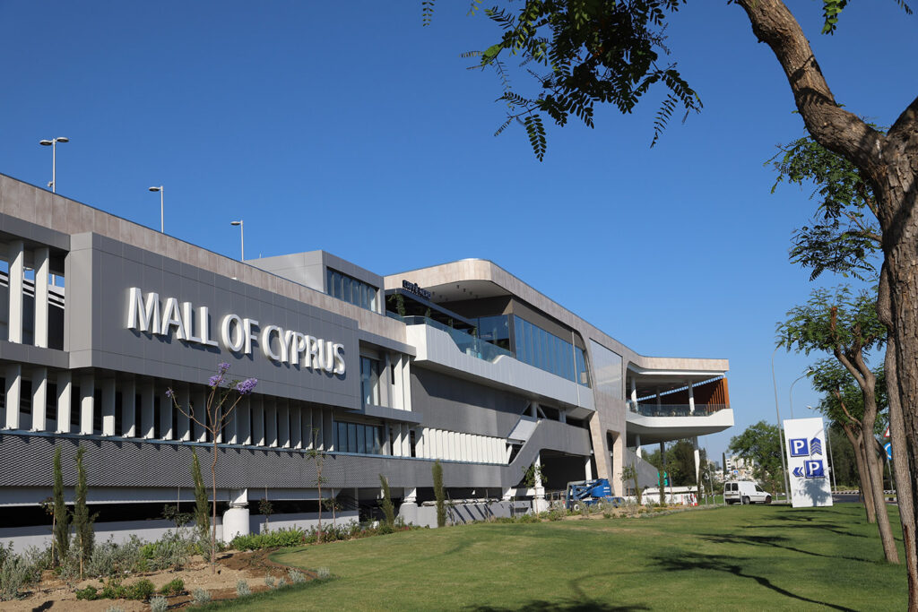 Modern exterior view of Mall of Cyprus, a standout among Pareto portfolio shopping centres, with large signage, green lawn, and parking signs visible under a clear blue sky. - Pareto Properties