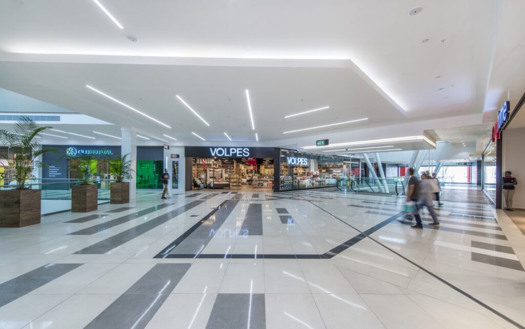 Wide view of a modern shopping centre corridor with polished floors, bright lighting, and several retail shops, including Volpes; a few blurred shoppers walk by. - Pareto Properties