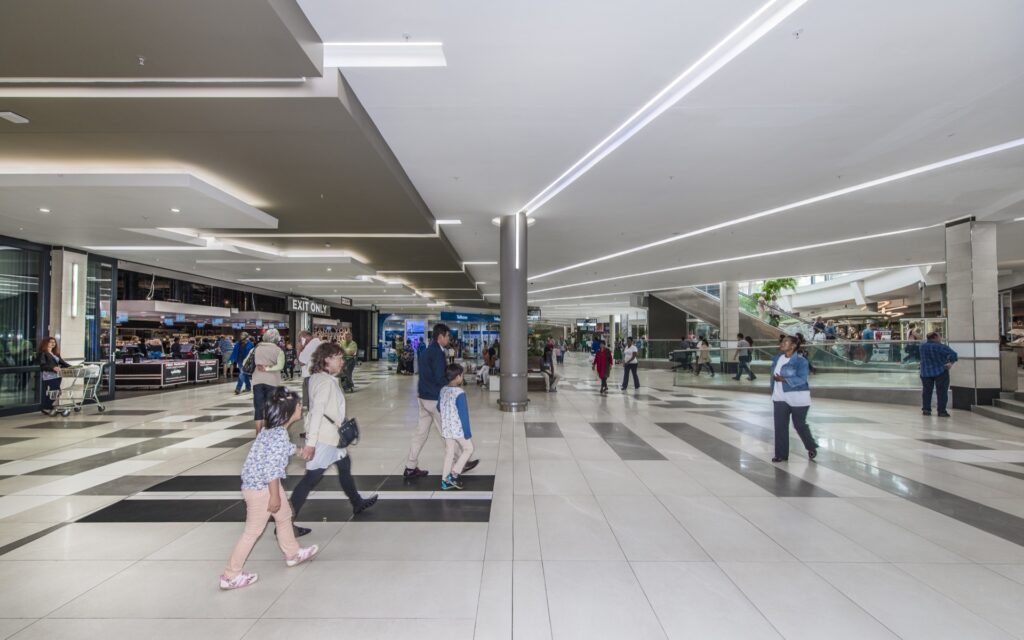 People walk through a spacious, modern shopping centre with tiled floors, bright lighting, and various shops and cafÃ©s visible in the background. - Pareto Properties