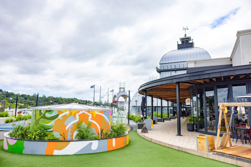 Outdoor dining area with a modern glass building, colourful painted planter, green artificial turf, and cloudy sky in the background. - Pareto Properties