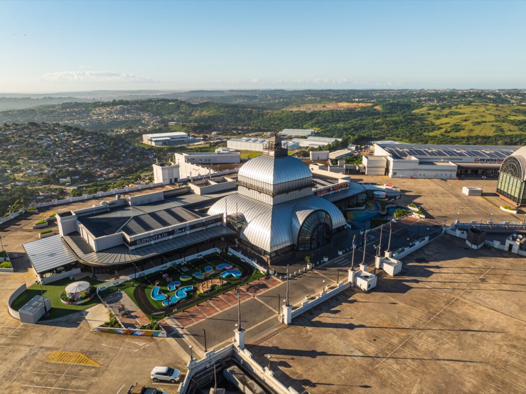 Aerial view of a large building complex with a domed central structure, outdoor play area, car park, and surrounding green hills under a clear sky. - Pareto Properties