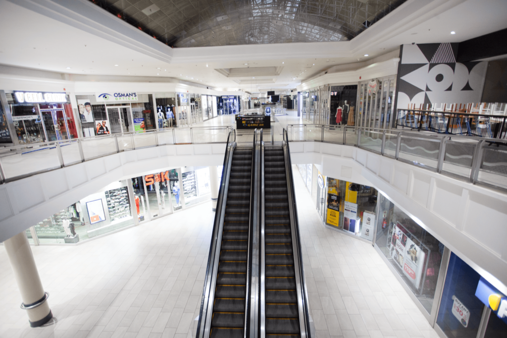 A wide, empty escalator in the centre of a bright, modern shopping centre with various closed shops on both levels. - Pareto Properties