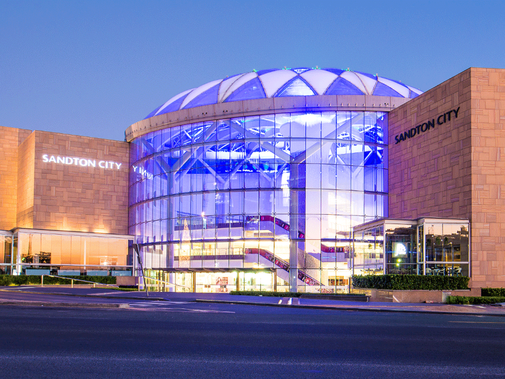 Sandton City shopping centre exterior, part of the distinguished Pareto portfolio shopping centres, features a glass dome, tan stone faÃ§ade, and illuminated blue and white lights at twilight. - Pareto Properties